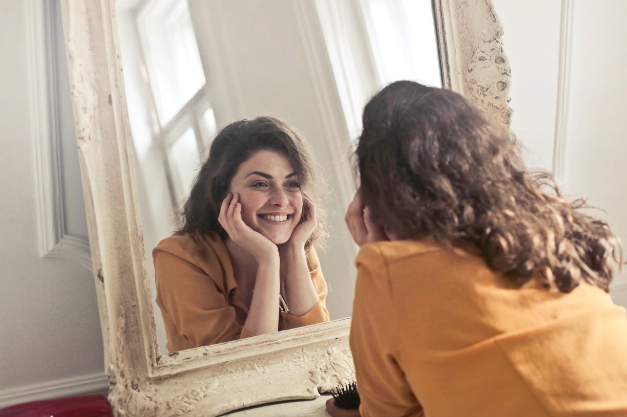 embark A cheerful woman smiles at her reflection in a vintage-style mirror, exuding positivity and warmth.